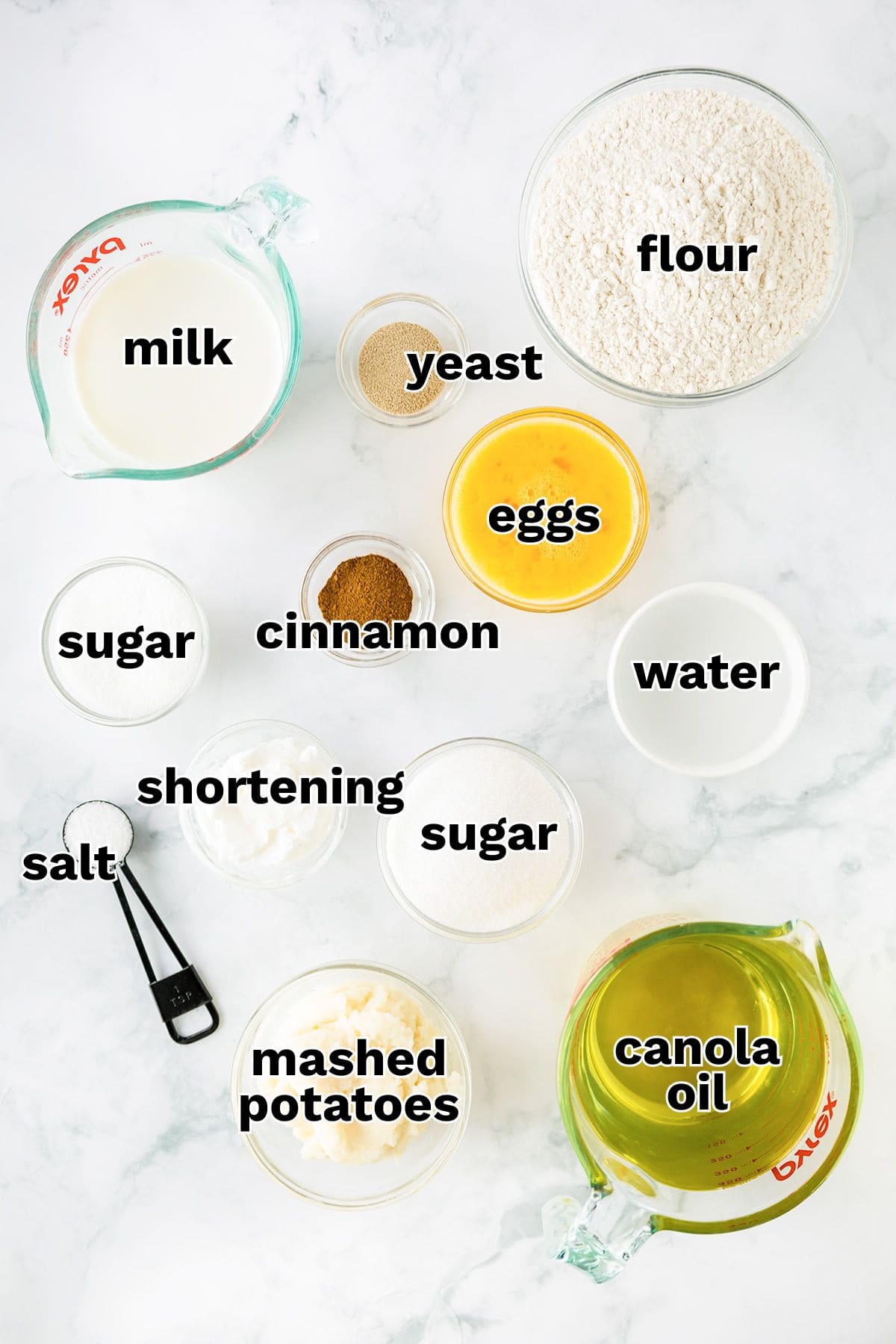 Top-down view of labeled baking ingredients for Potato Donuts on a marble surface, including flour, milk, yeast, eggs, water, sugar, cinnamon, salt, shortening, mashed potatoes, and canola oil.