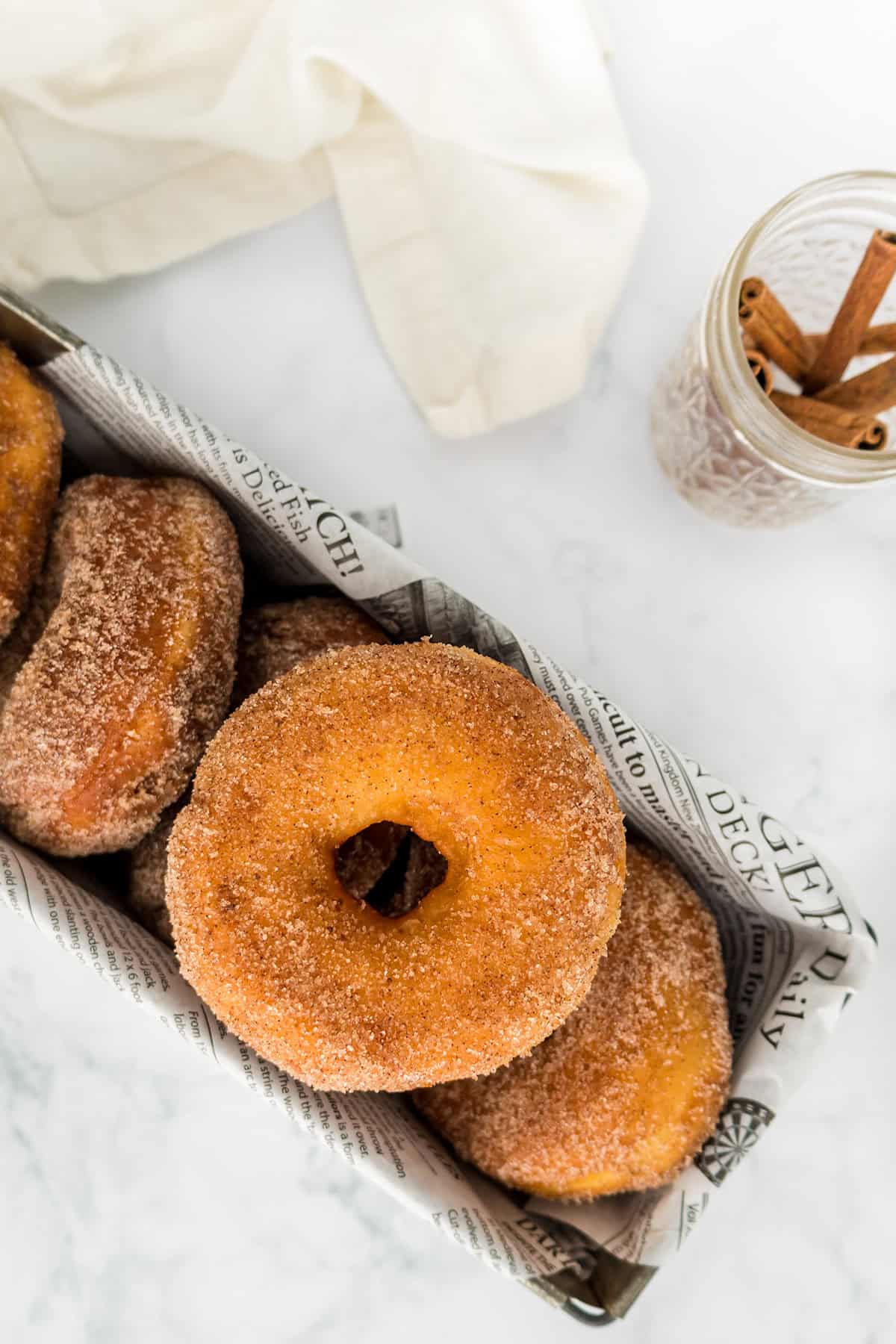 A basket lined with newspaper holds several cinnamon sugar Potato Donuts. A glass jar with cinnamon sticks and a white cloth are in the background on a marble surface.