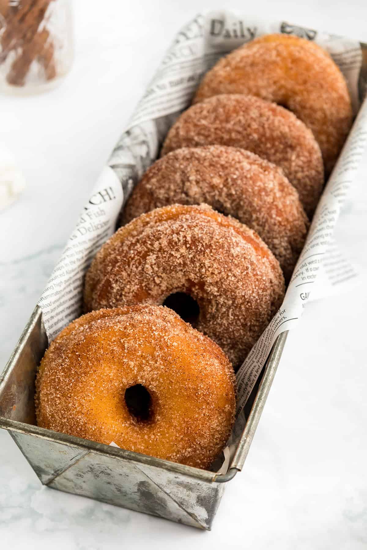 Six cinnamon sugar potato donuts are arranged upright in a metal tray lined with newspaper print parchment paper on a light surface.