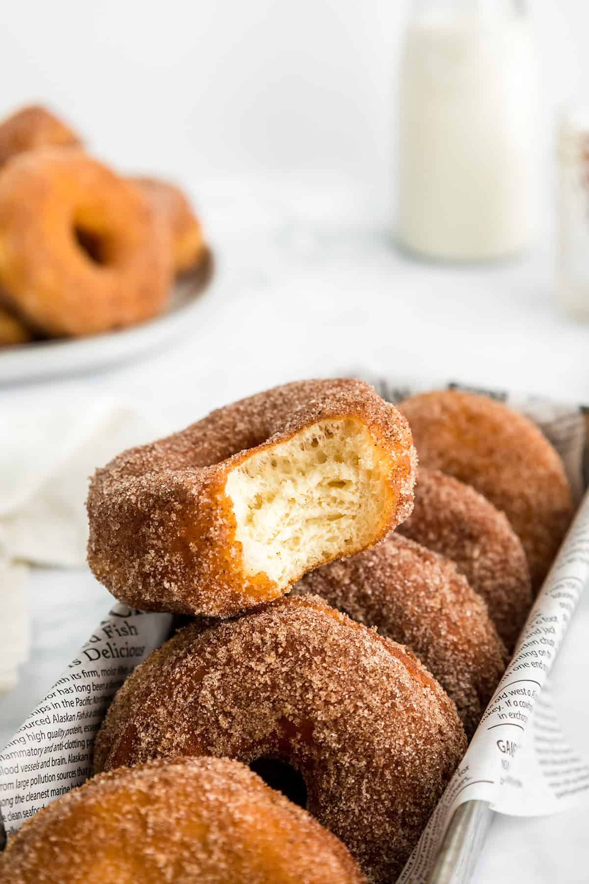 Tray of cinnamon sugar potato donuts with one donut showing a bite taken out, placed on newspaper, with a blurred plate of donuts and a milk bottle in the background.
