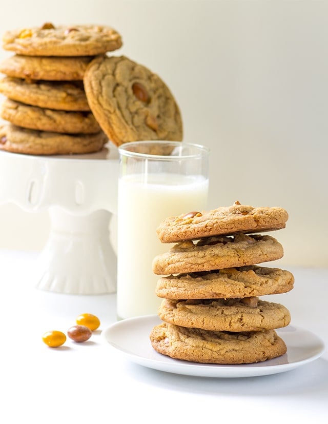 a white plate stacked high with butterscotch M&M cookies next to a glass of milk