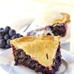 A slice of blueberry pie on a plate with a glass of milk and Concord Grape Pie.