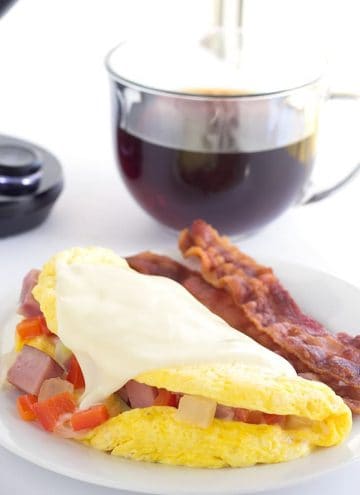 western omelet and bacon on a white plate with a cup of coffee behind it