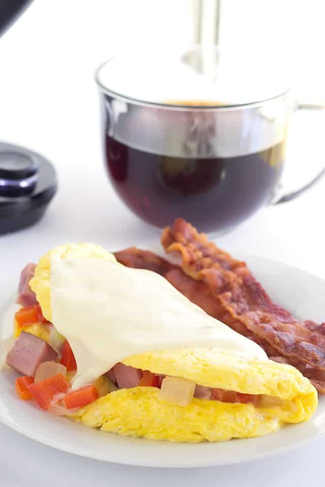 western omelet and bacon on a white plate with a cup of coffee behind it