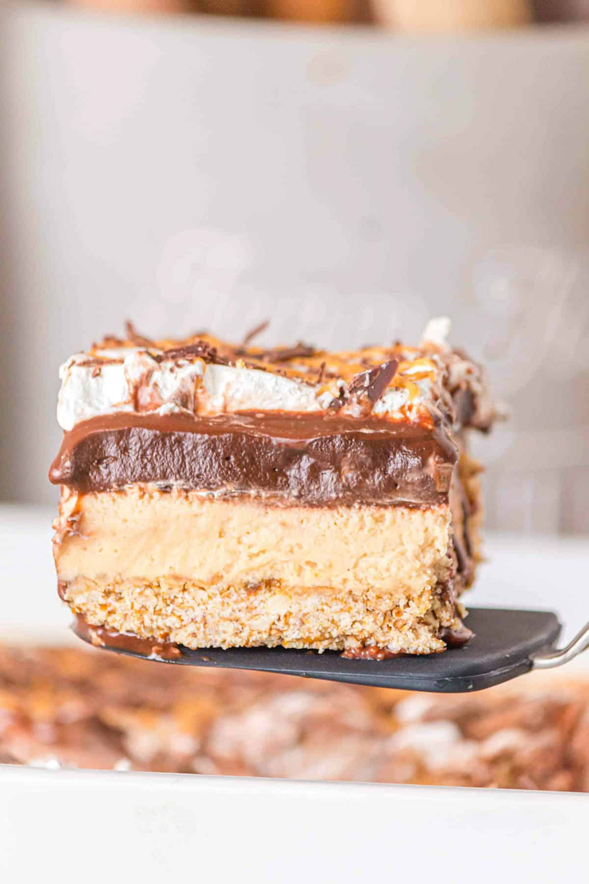 A close-up of a Chocolate Peanut Butter Layer Dessert square with a graham cracker crust, creamy filling, chocolate layer, and whipped topping, held up on a spatula.