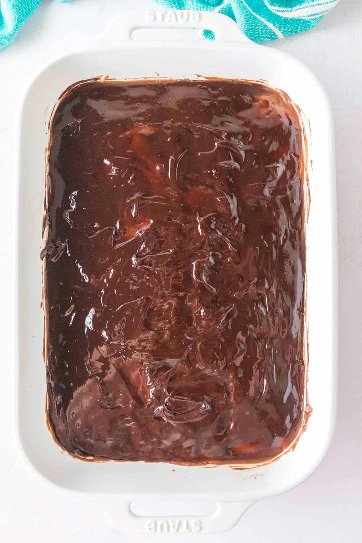 A white rectangular baking dish filled with a rich chocolate dessert, possibly brownies or cake, topped with a glossy layer of frosting, reminiscent of a decadent Chocolate Peanut Butter Layer Dessert.