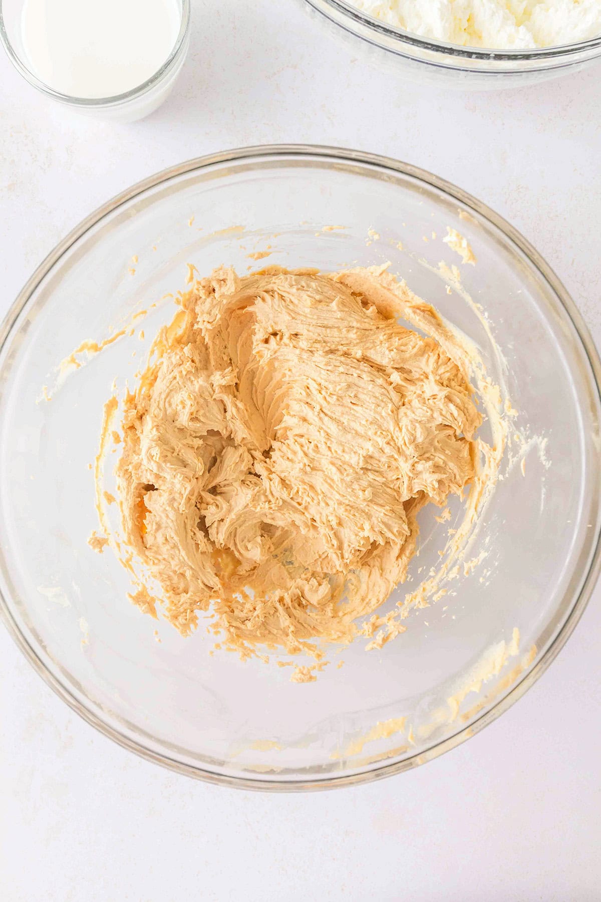 A glass bowl containing creamed butter and sugar mixture sits on a white surface, ready for the next step in creating a decadent Chocolate Peanut Butter Layer Dessert, with part of a milk glass visible nearby.
