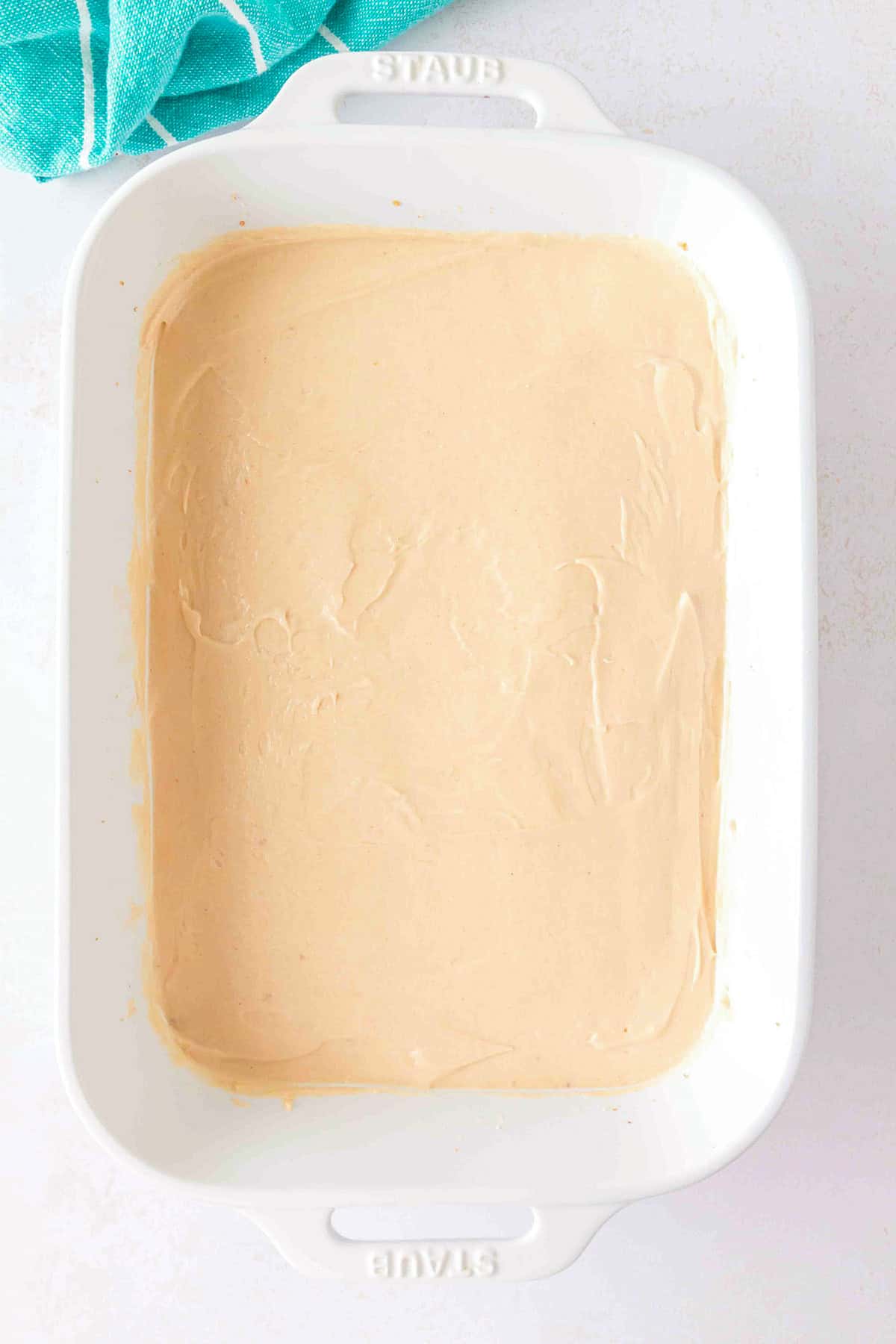 A rectangular white baking dish contains an even layer of beige batter for a Chocolate Peanut Butter Layer Dessert, with a blue towel partially visible in the top left corner.