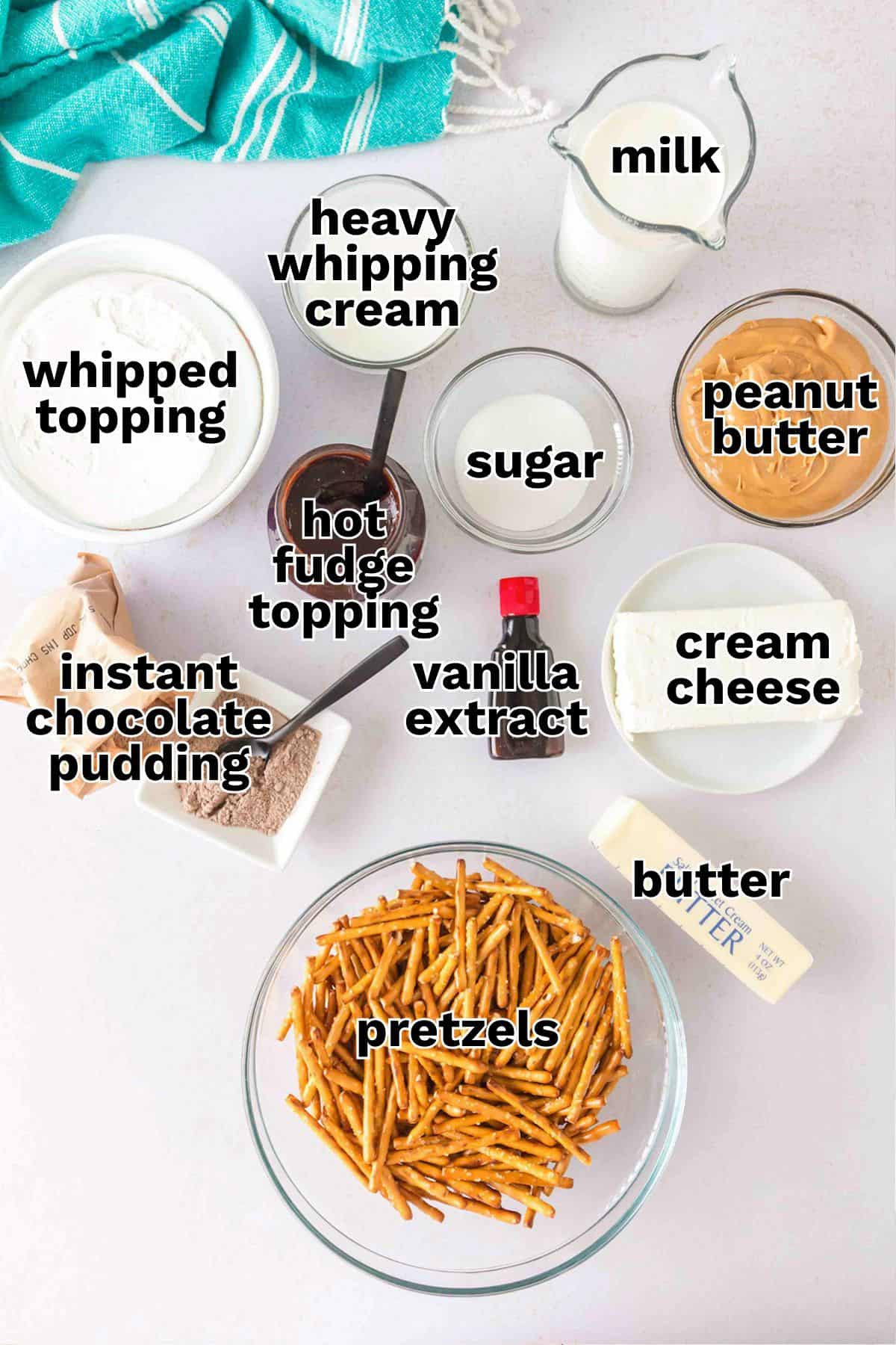 A top-down view of ingredients in bowls and containers for a Chocolate Peanut Butter Layer Dessert recipe, labeled: whipped topping, instant chocolate pudding, pretzels, butter, cream cheese, peanut butter, sugar, milk, heavy whipping cream, hot fudge topping, and vanilla extract.