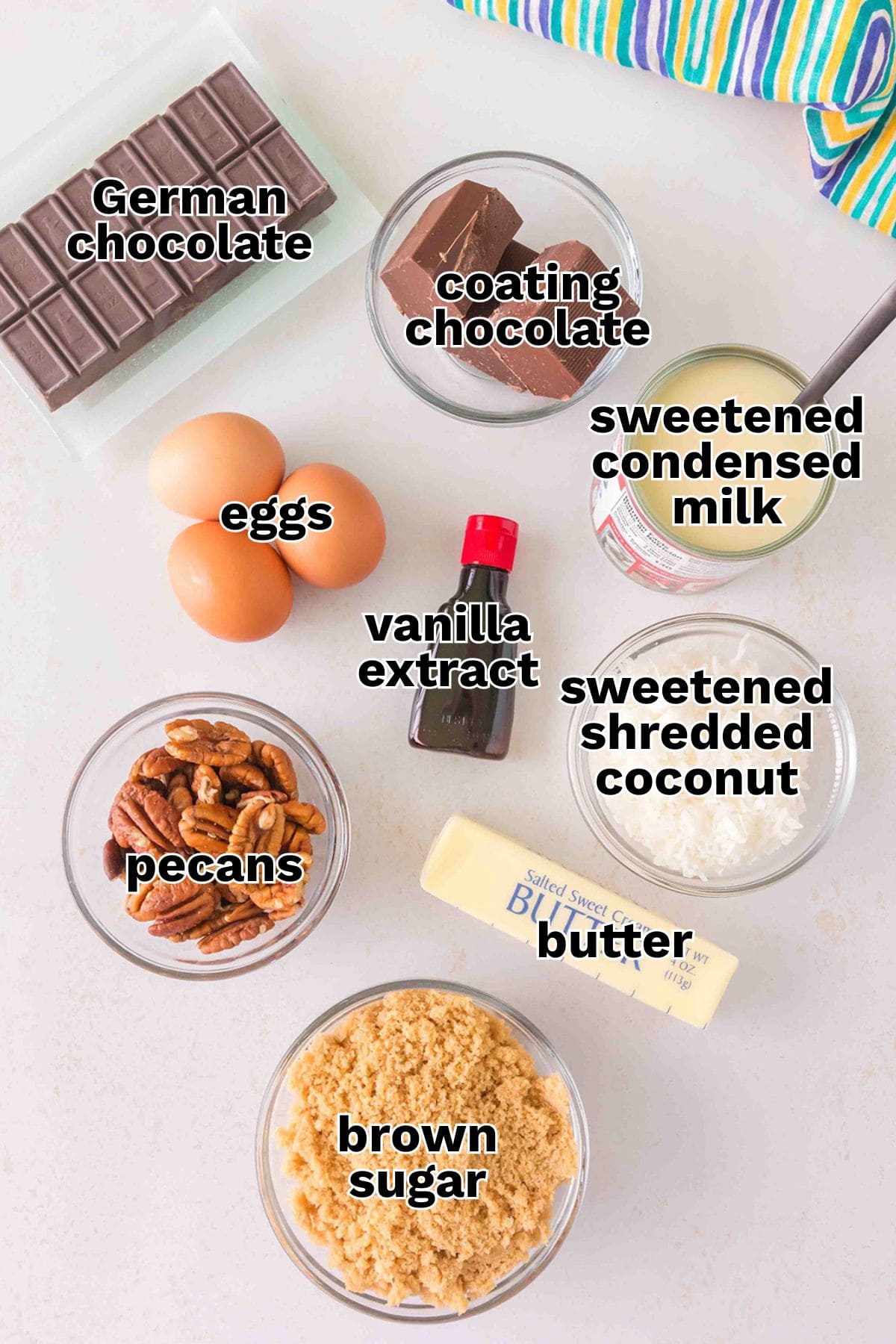 Ingredients for baking are arranged on a table, ready for making German Chocolate Truffles, including German chocolate, coating chocolate, eggs, sweetened condensed milk, vanilla extract, pecans, butter, brown sugar, and shredded coconut.