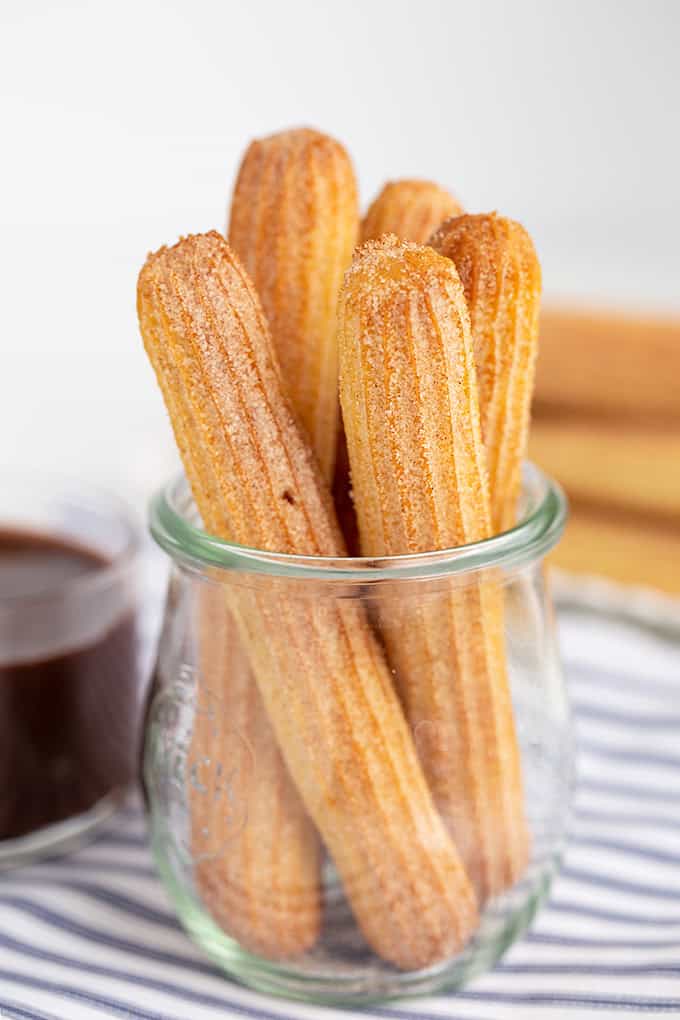 Several baked churros coated in cinnamon sugar are arranged upright in a glass jar, with a small dish of chocolate sauce and a striped cloth in the background.