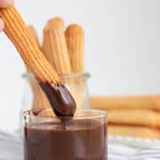 A hand dips a baked churro into a glass of chocolate sauce, with more baked churros in a glass jar and on a plate in the background.