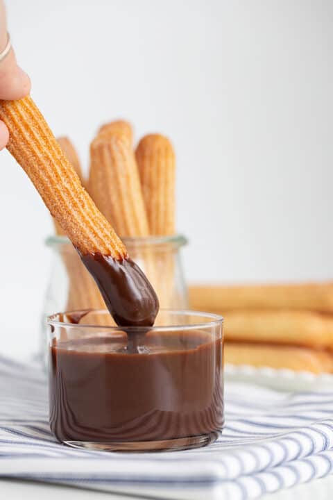 A hand dips a baked churro into a glass of chocolate sauce, with more baked churros in a glass jar and on a plate in the background.