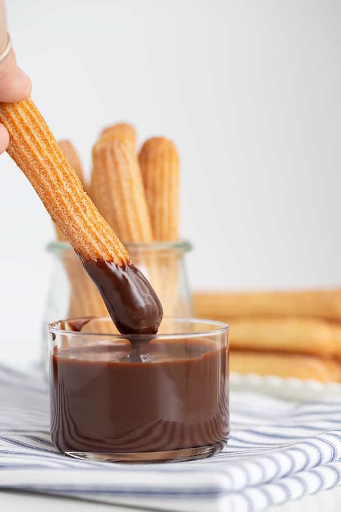 A hand dips a baked churro into a glass of chocolate sauce, with more baked churros in a glass jar and on a plate in the background.