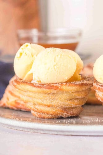 A Churro Ice Cream Cup dusted with sugar is topped with two scoops of vanilla ice cream, served on a gray plate.