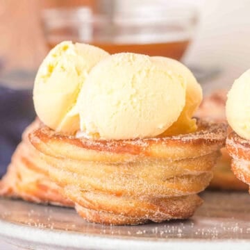 A Churro Ice Cream Cup dusted with sugar is topped with two scoops of vanilla ice cream, served on a gray plate.