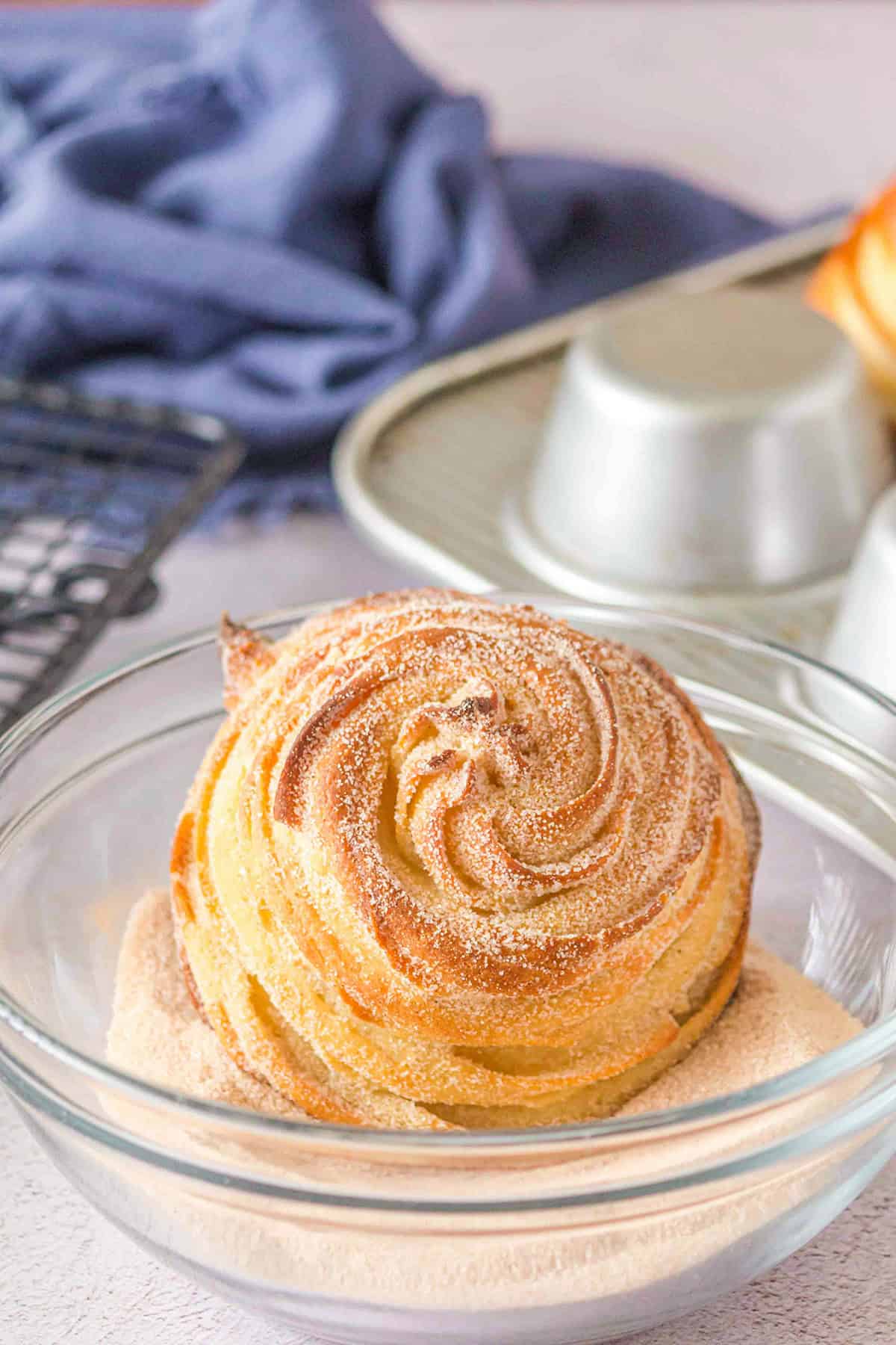 A swirled pastry coated with cinnamon sugar sits in a clear glass bowl, resembling delicious Churro Ice Cream Cups, with a metal baking pan and a blue cloth in the background.