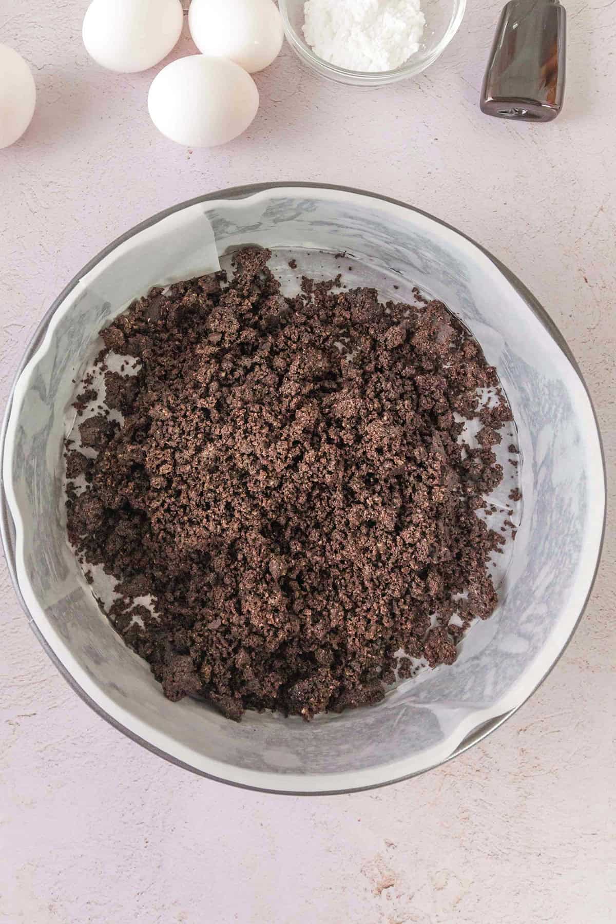 A mixing bowl filled with crushed chocolate cookie crumbs, perfect for the base of a Triple Chocolate Cheesecake, with eggs, a small bowl of white powder, and a bottle of vanilla extract visible nearby.