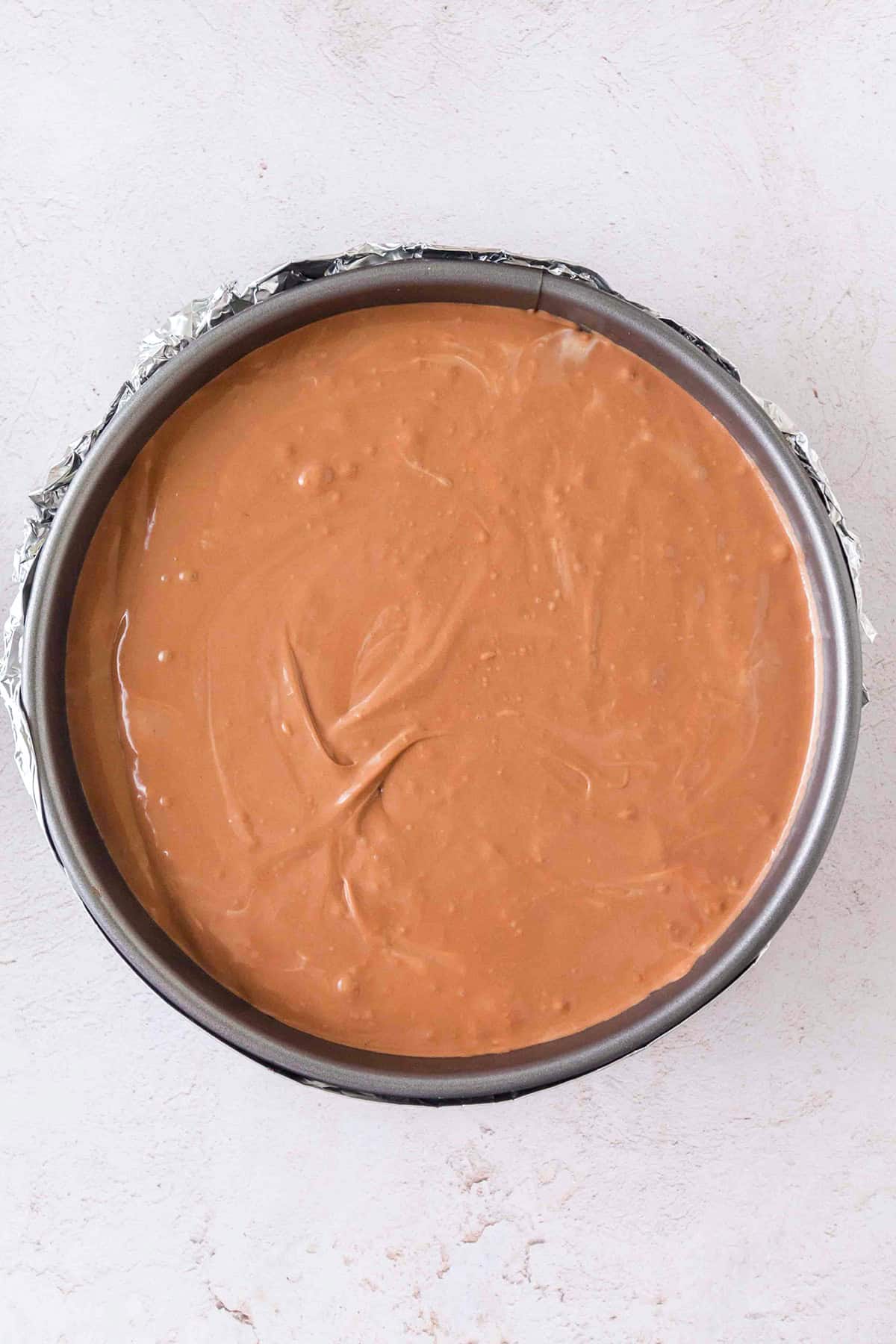 A round baking pan lined with foil is filled with smooth, light brown Triple Chocolate Cheesecake batter, ready to be baked; the surface has some visible swirls.