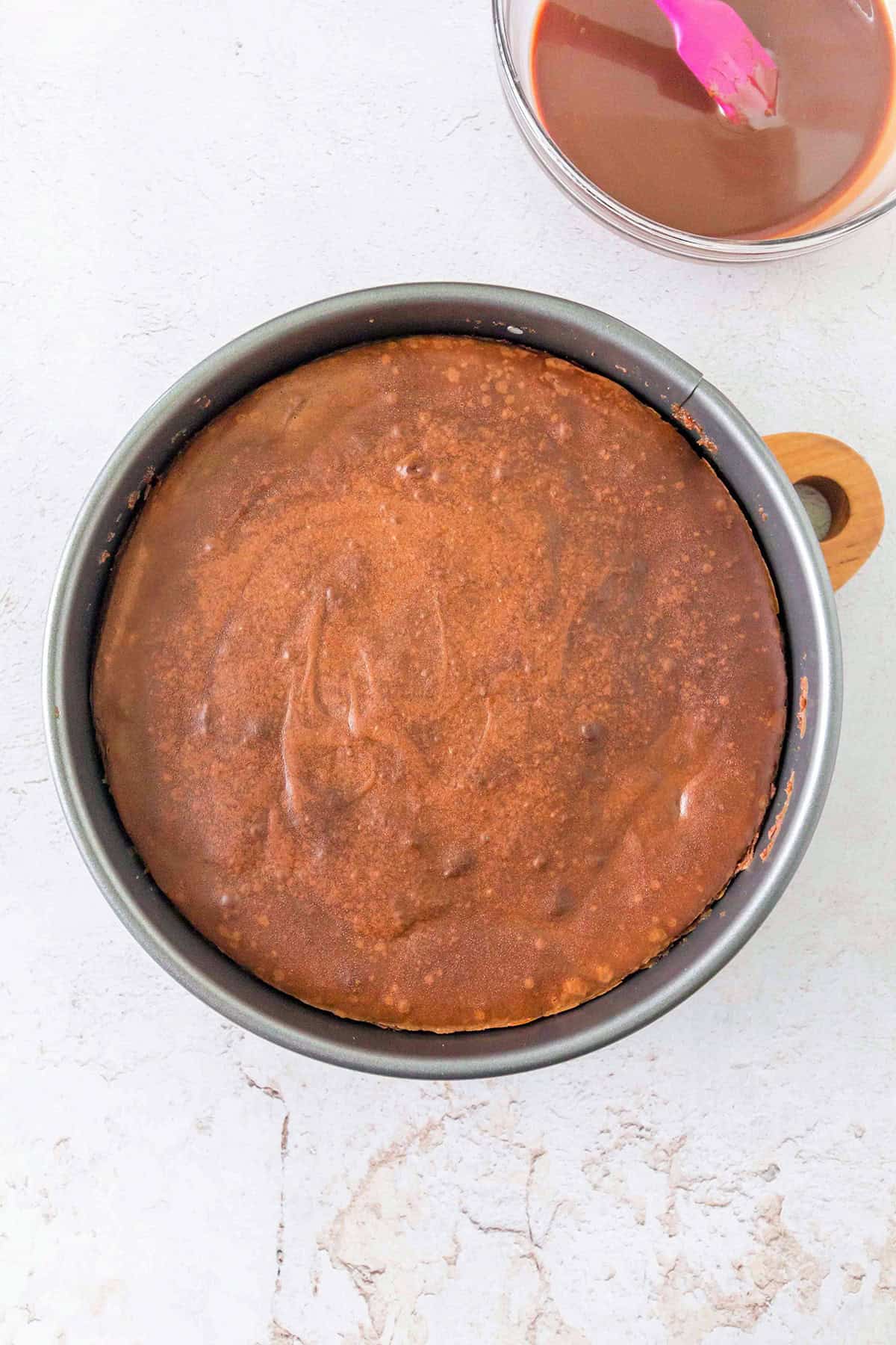 A round Triple Chocolate Cheesecake in a metal pan sits on a light countertop. Nearby, a bowl with chocolate glaze and a pink spatula is partially visible.