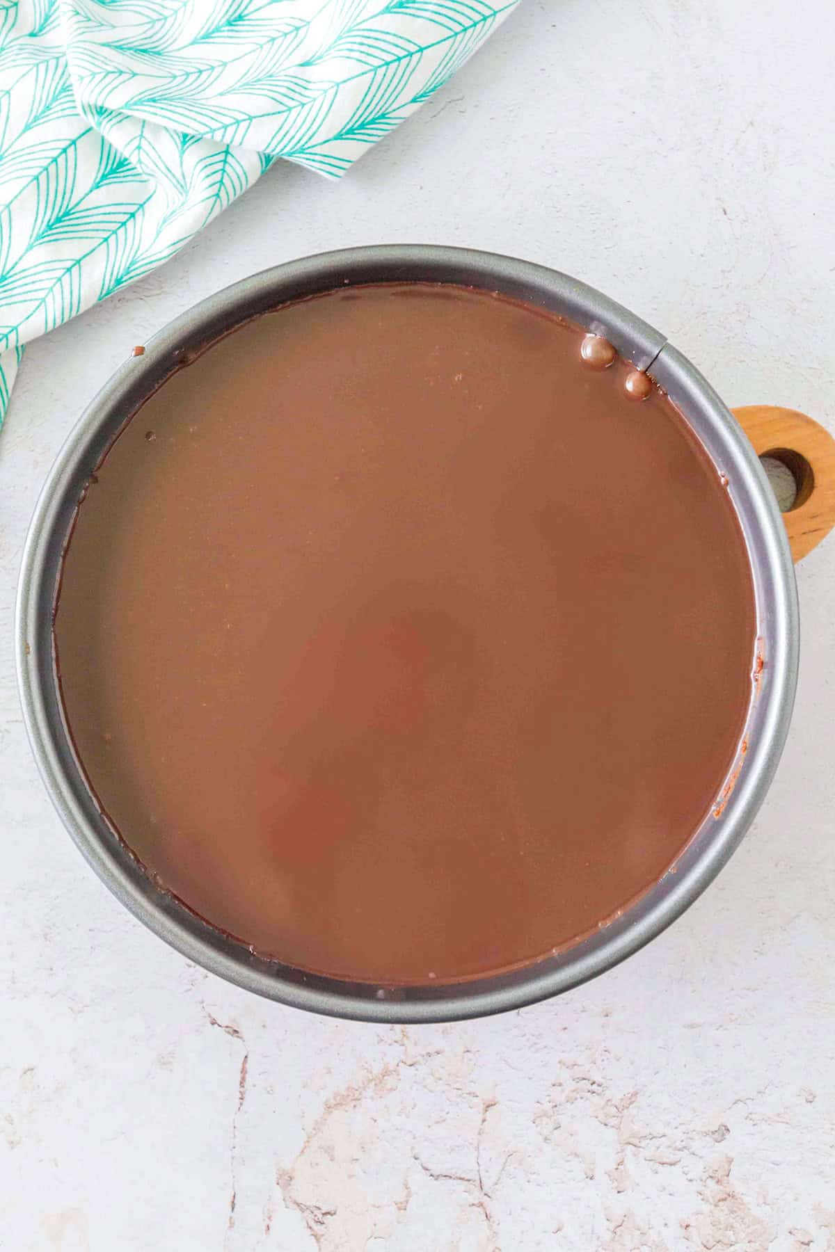 A round cake pan filled with smooth, glossy chocolate ganache rests on a light surface near a green and white patterned cloth, promising the rich indulgence of a Triple Chocolate Cheesecake.