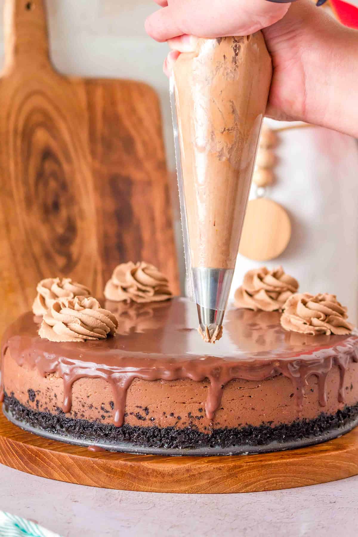 A hand is piping chocolate frosting onto a Triple Chocolate Cheesecake with an Oreo crust and chocolate ganache, which sits on a wooden board.