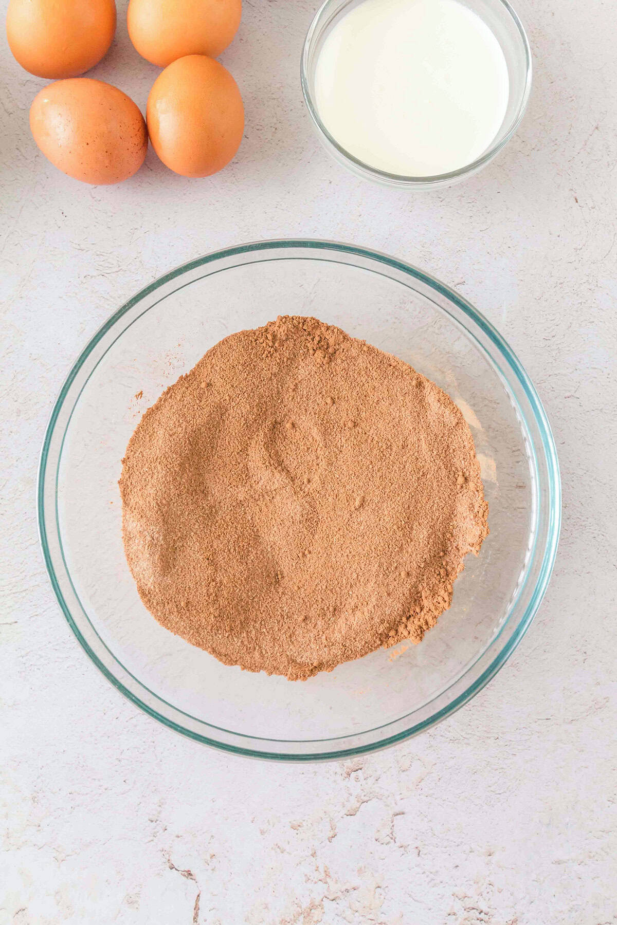 A glass bowl filled with cocoa powder, next to three eggs and a glass bowl of milk on a light-colored countertop—the perfect beginning for crafting a decadent Triple Chocolate Cheesecake.
