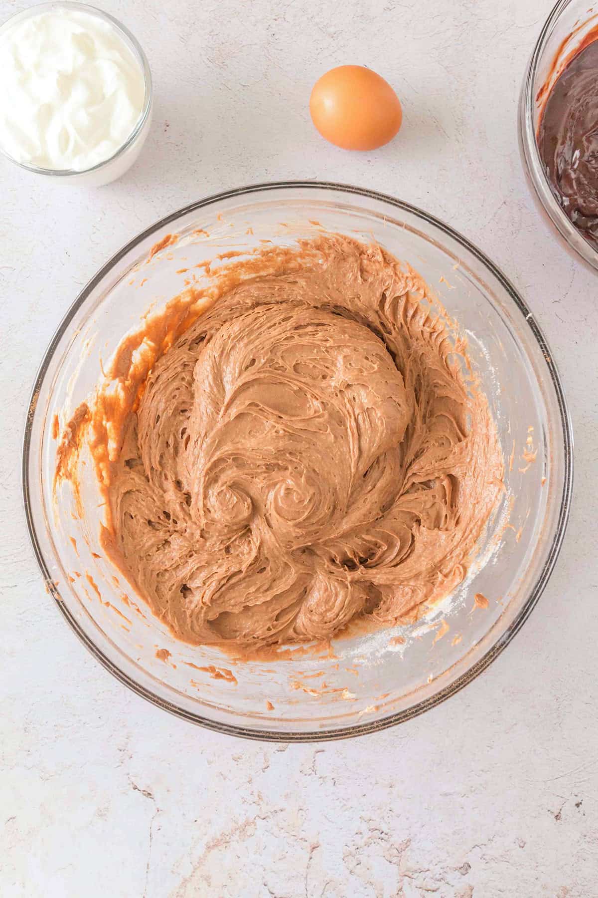 A glass bowl filled with light brown cake batter sits on a countertop next to an egg, a bowl of yogurt, and a bowl of chocolate—perfect ingredients for preparing a decadent Triple Chocolate Cheesecake.