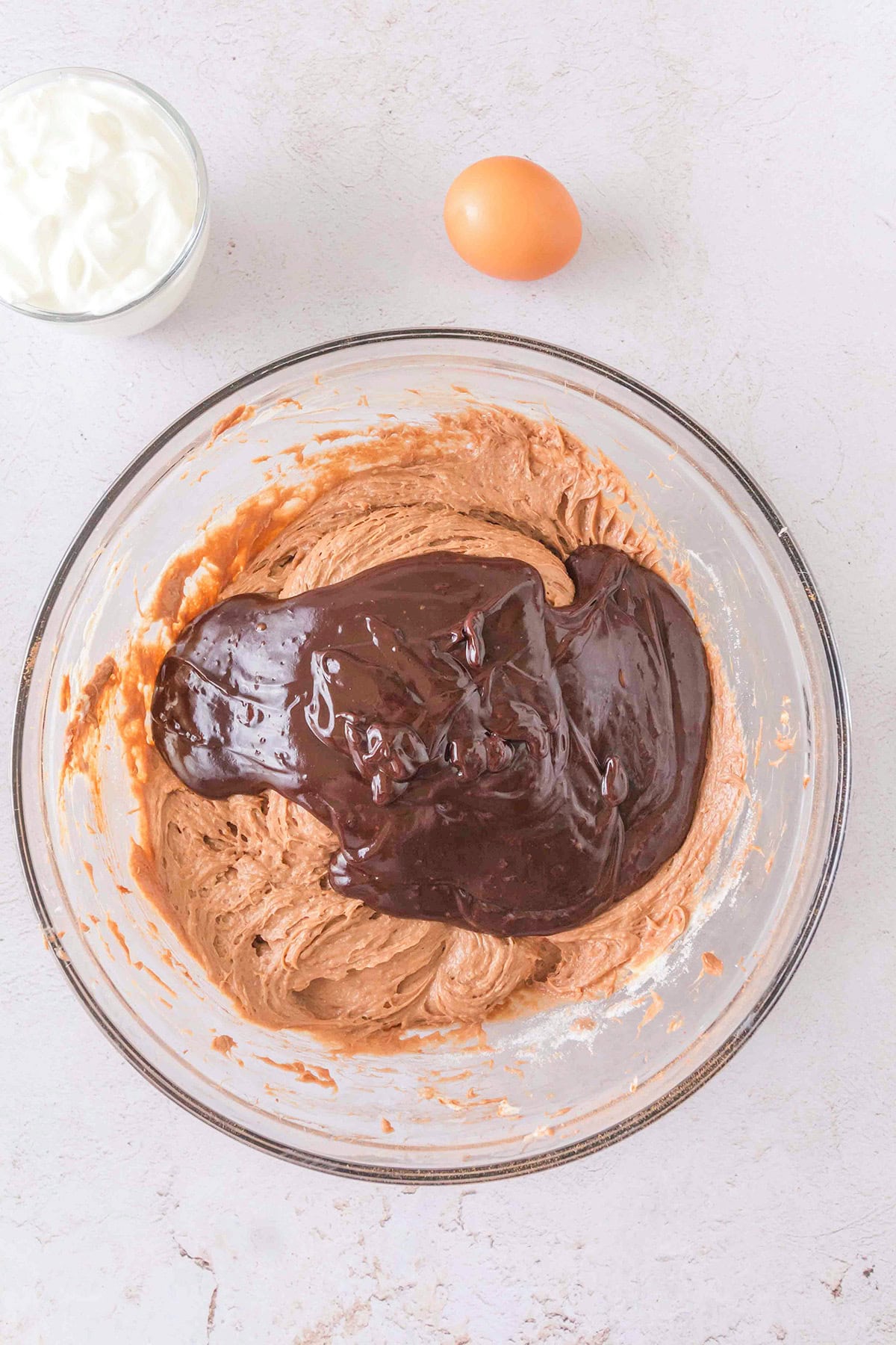 A glass bowl with chocolate batter being mixed for Triple Chocolate Cheesecake, next to a raw egg and a small bowl of white yogurt on a light surface.