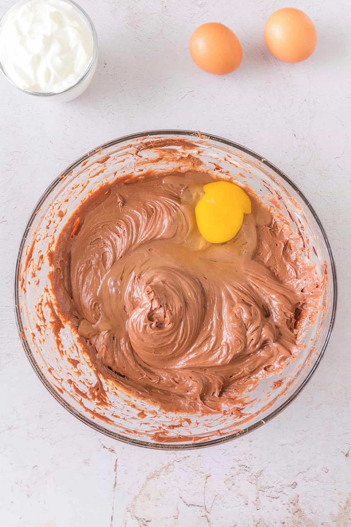 A glass bowl of chocolate batter, perfect for a decadent Triple Chocolate Cheesecake, with one cracked egg on top, sits next to two whole eggs and a small bowl of white cream on a light surface.