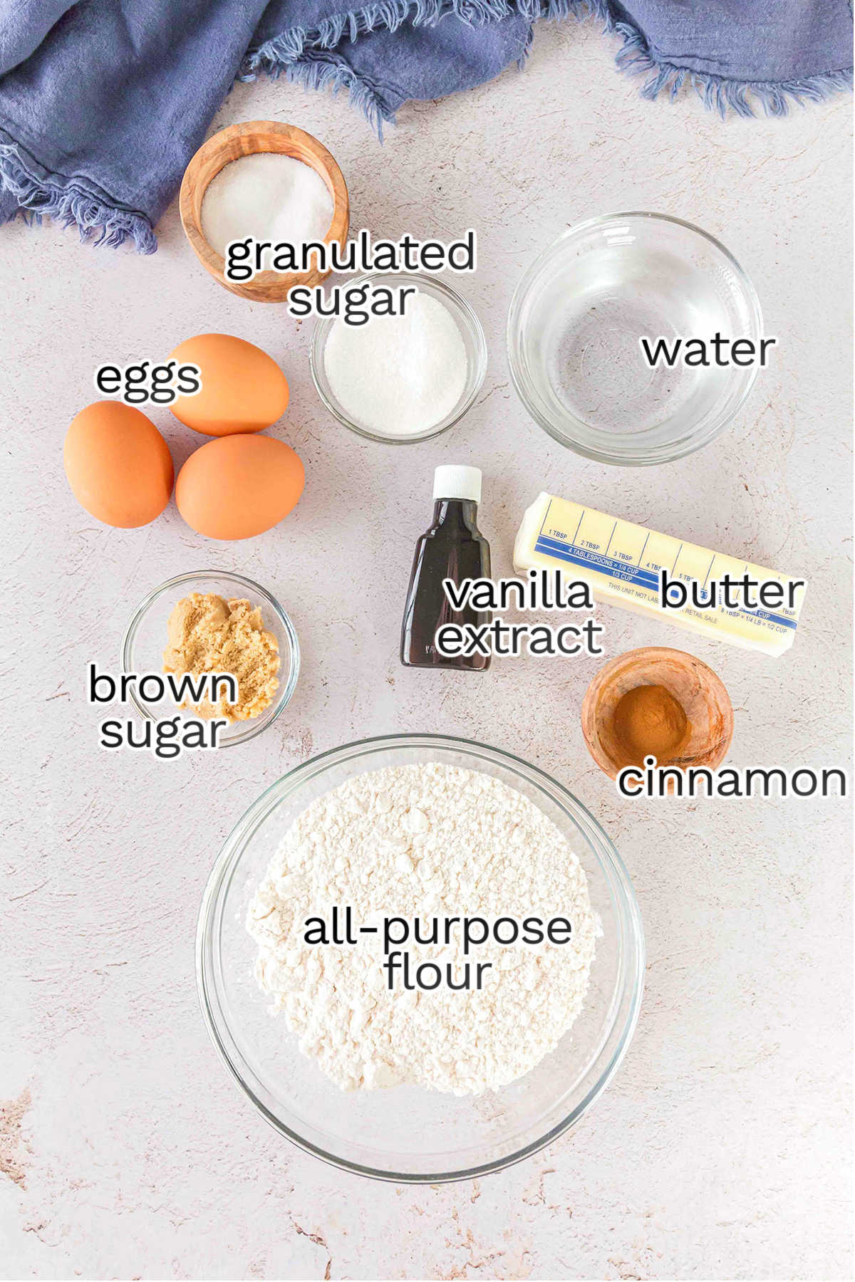 A flat lay of baking ingredients for Churro Ice Cream Cups, including eggs, granulated sugar, brown sugar, water, vanilla extract, butter, cinnamon, and all-purpose flour—all clearly labeled.