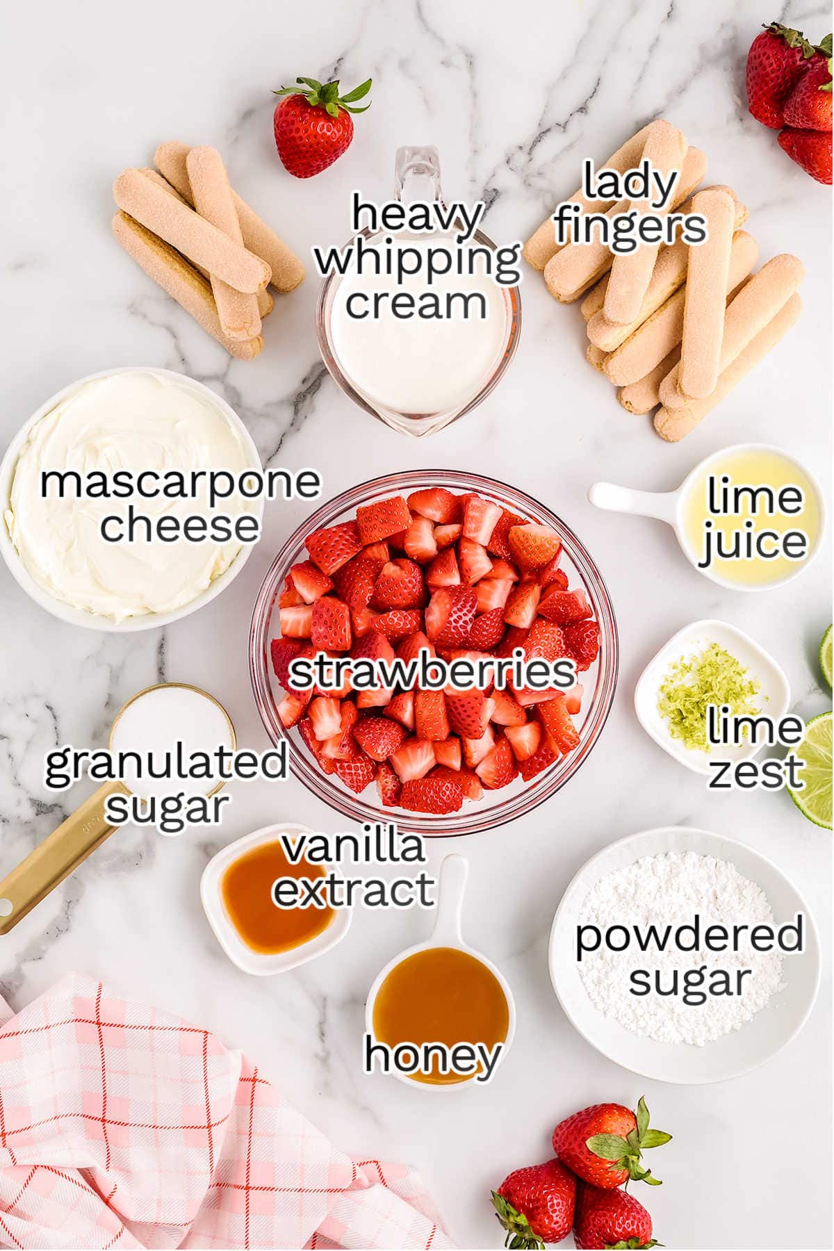 A top-down view of ingredients for a strawberry tiramisu dessert, including strawberries, ladyfingers, mascarpone cheese, heavy whipping cream, granulated and powdered sugar, honey, lime juice, lime zest, and vanilla extract.