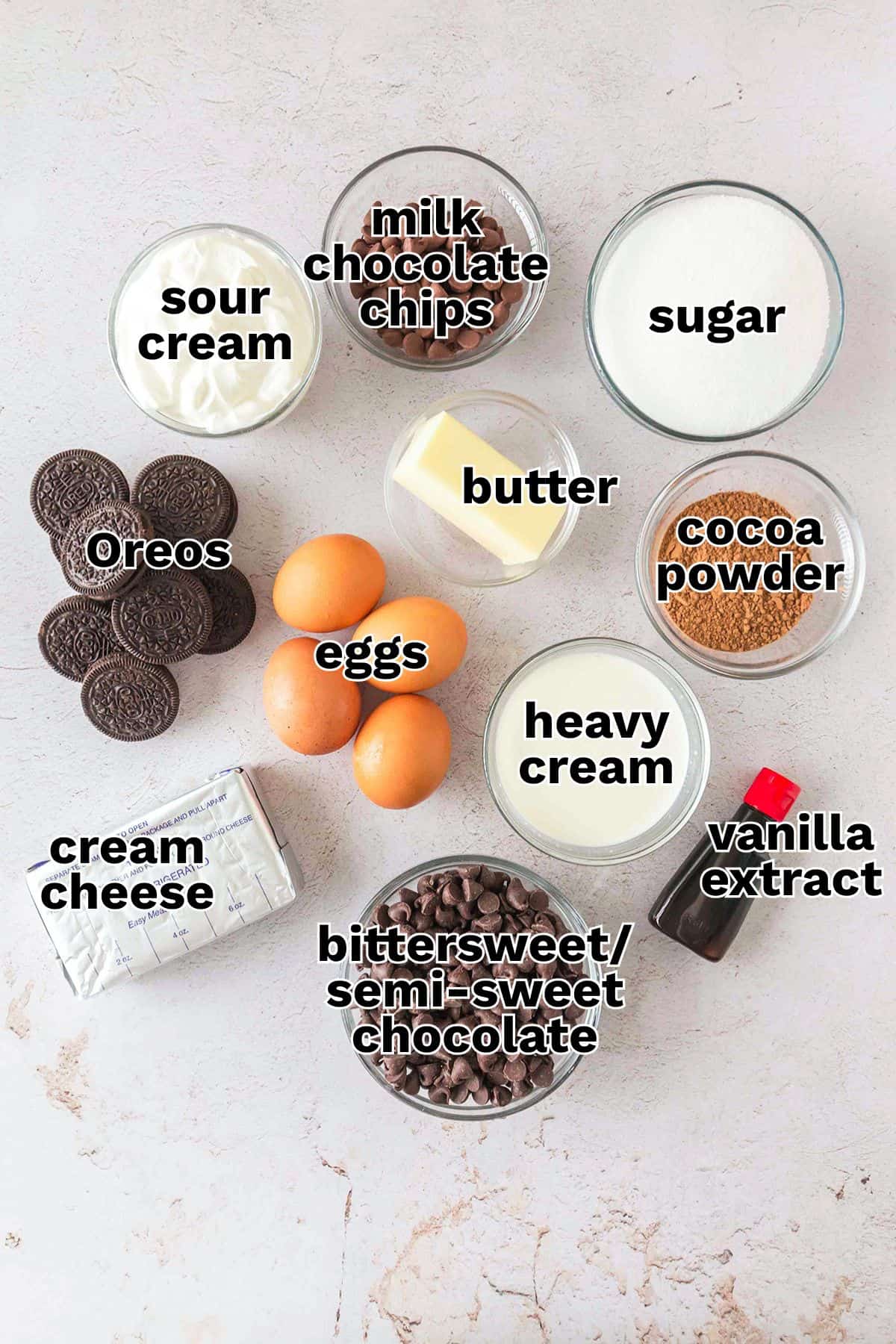 Top-down view of labeled ingredients in bowls and on a table for a decadent Triple Chocolate Cheesecake, including Oreos, cream cheese, eggs, sour cream, milk chocolate chips, sugar, butter, cocoa powder, heavy cream, vanilla, and chocolate chips.