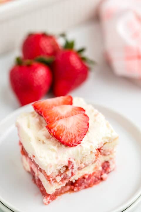 A slice of layered strawberry tiramisu topped with whipped cream and two sliced strawberries, served on a white plate with whole strawberries in the background.