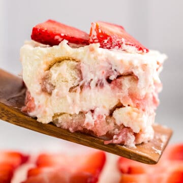 A close-up of a slice of strawberry tiramisu dessert with creamy layers and sliced strawberries on top, served on a wooden spatula.