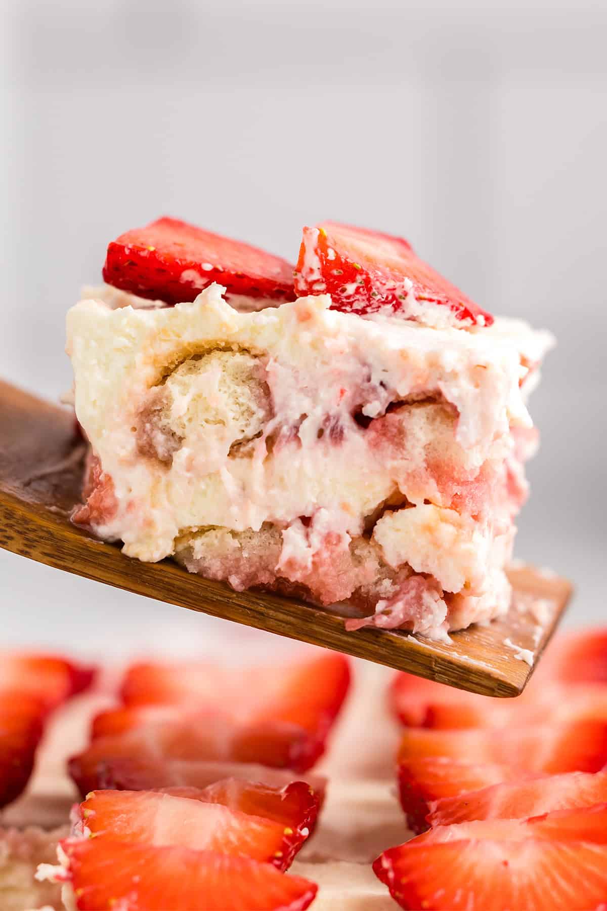 A close-up of a slice of strawberry tiramisu dessert with creamy layers and sliced strawberries on top, served on a wooden spatula.