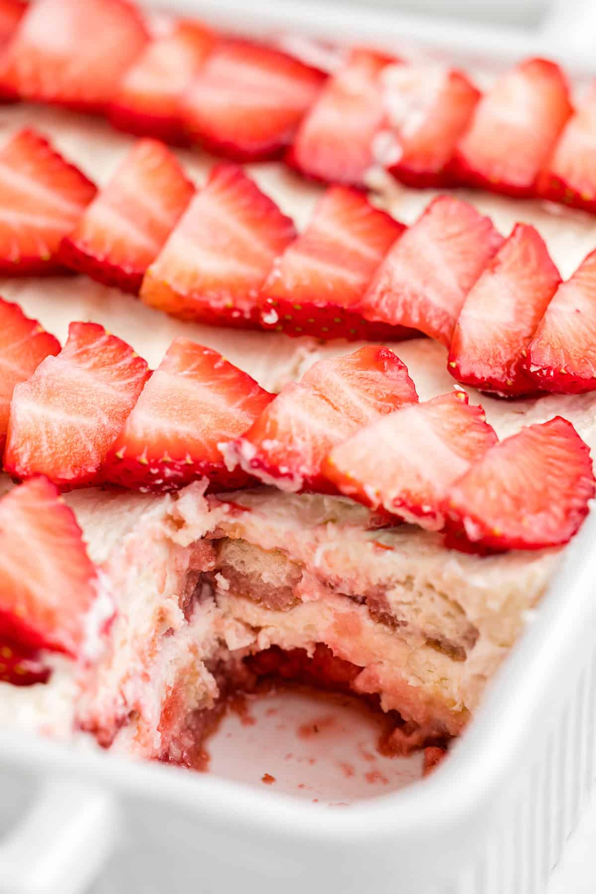 A close-up of a partially eaten strawberry tiramisu in a white dish, topped with sliced strawberries and cream, with delicious layers visible underneath.