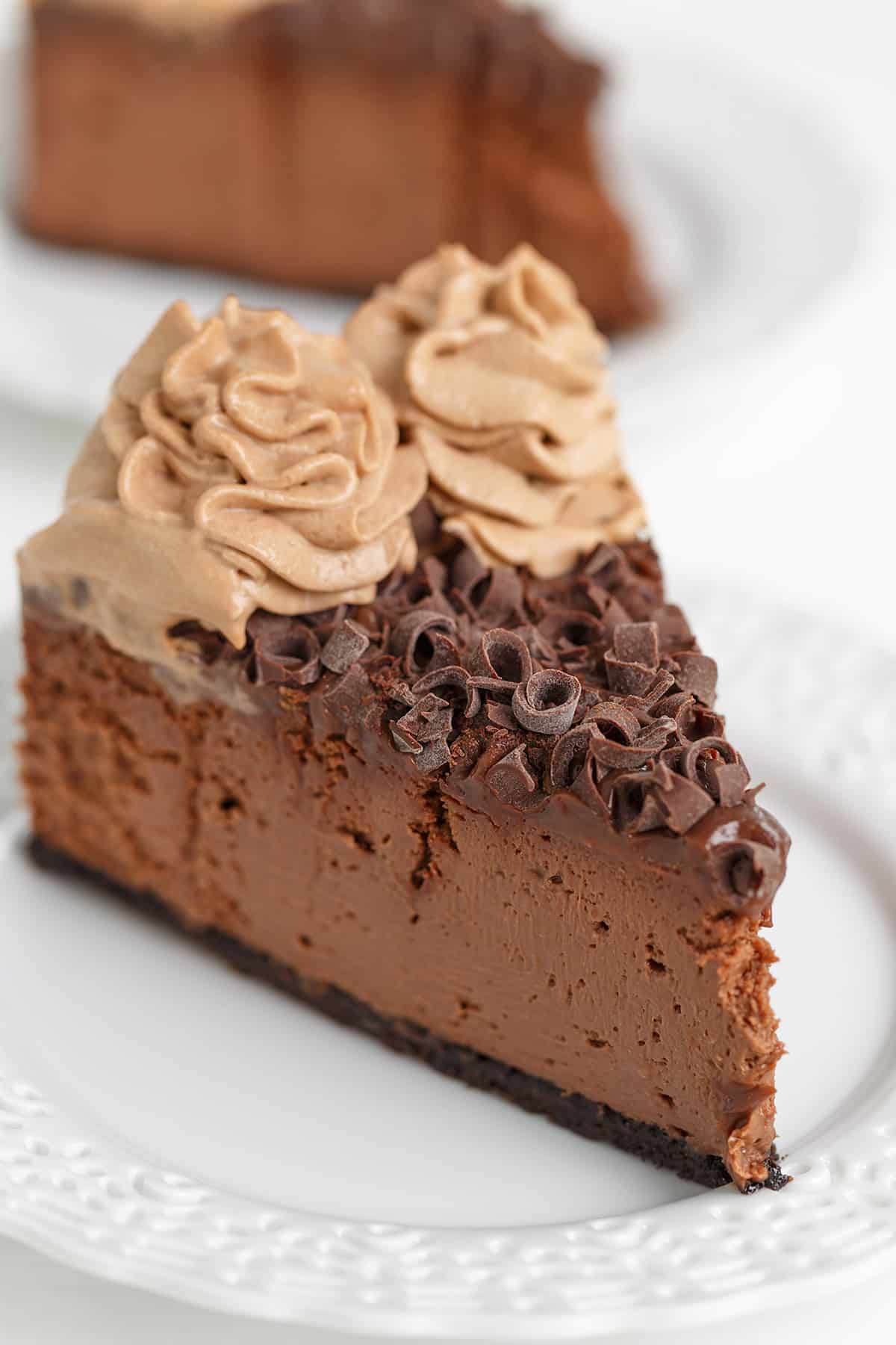 A slice of Triple Chocolate Cheesecake sits elegantly on a white plate, topped with chocolate shavings and swirls of chocolate whipped cream.