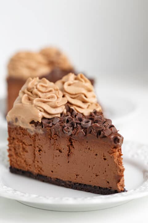 A slice of Triple Chocolate Cheesecake, topped with chocolate curls and swirls of chocolate whipped cream, sits on a white plate.