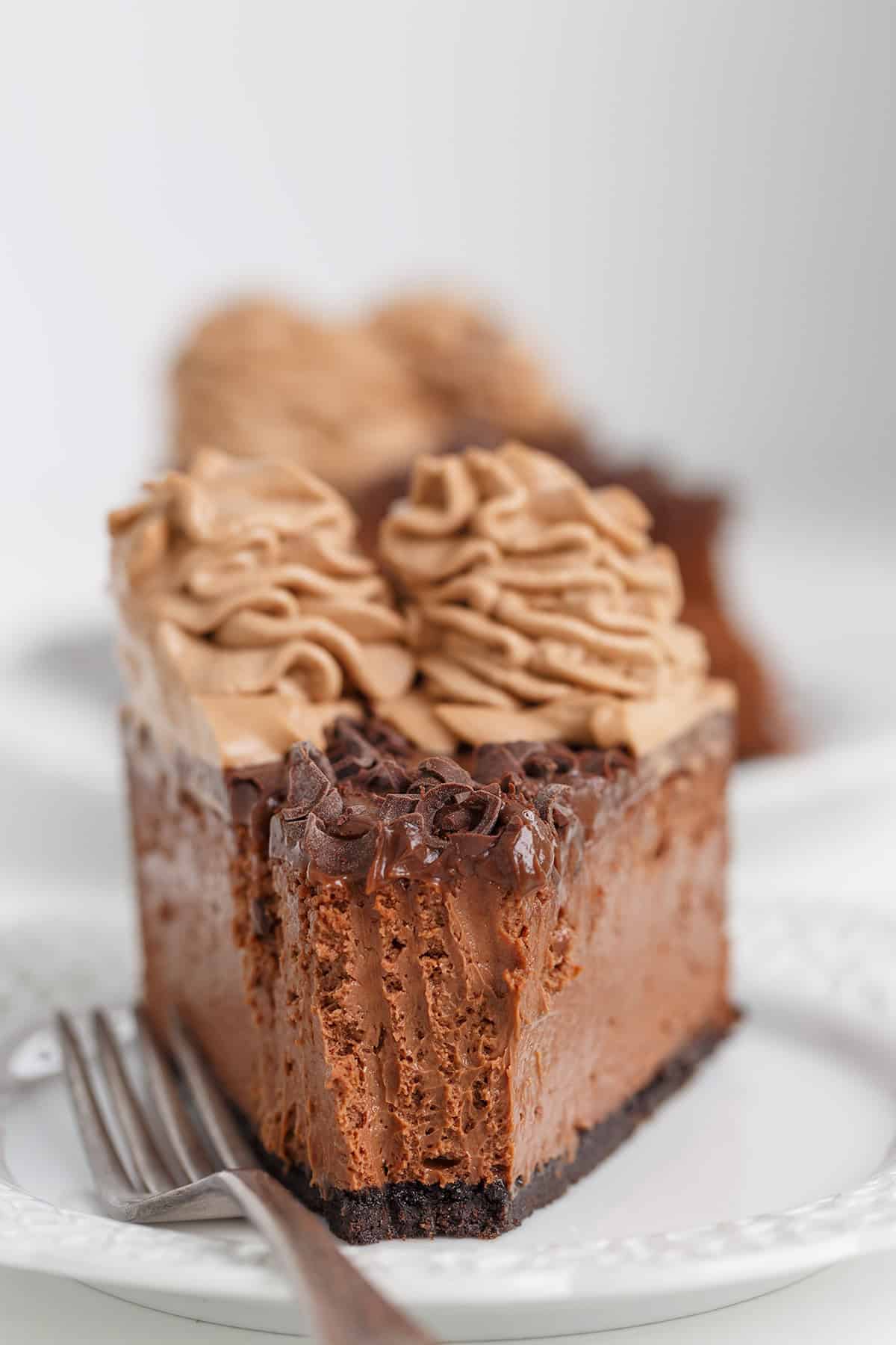 A slice of Triple Chocolate Cheesecake with whipped chocolate topping and a fork on a white plate, with a bite taken out.