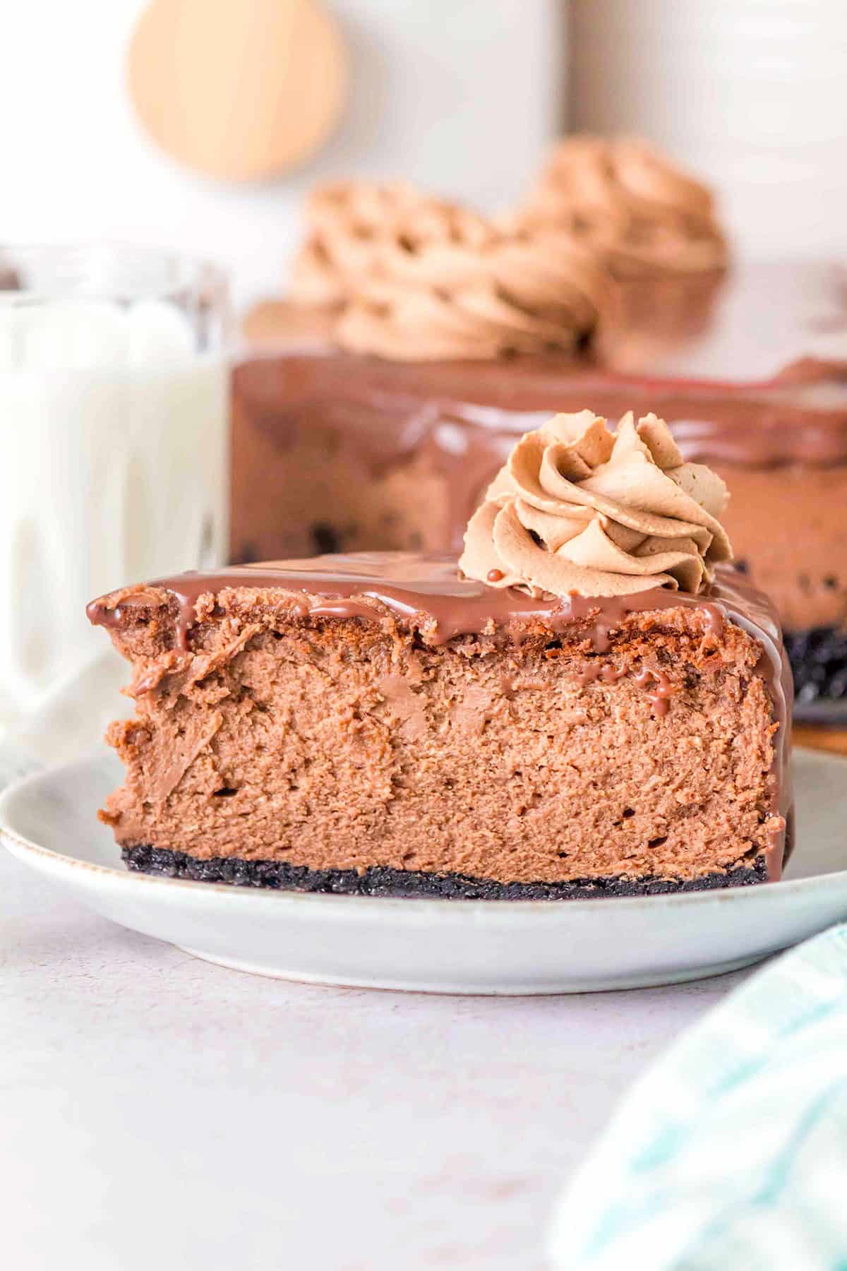 A slice of Triple Chocolate Cheesecake with rich chocolate frosting on top, placed on a white plate, is accompanied by a glass of milk and the rest of the decadent cake in the background.