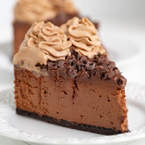 A slice of rich Triple Chocolate Cheesecake topped with chocolate shavings and swirls of chocolate mousse on a white plate.