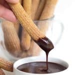 Baked Churros standing in a clear glass coffee mug. One churro being dipped into melted chocolate