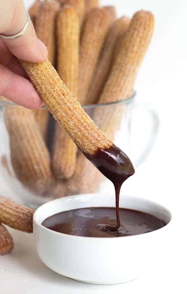 Baked Churros standing in a clear glass coffee mug. One churro being dipped into melted chocolate