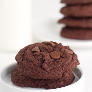 two double chocolate chip pudding cookies on a white plate with a stack of cookies in the background