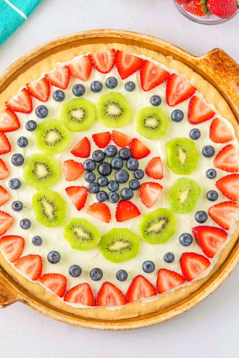 An easy fruit pizza with a cookie crust, cream cheese frosting, and toppings of sliced strawberries, kiwi, and blueberries arranged in a circular pattern.