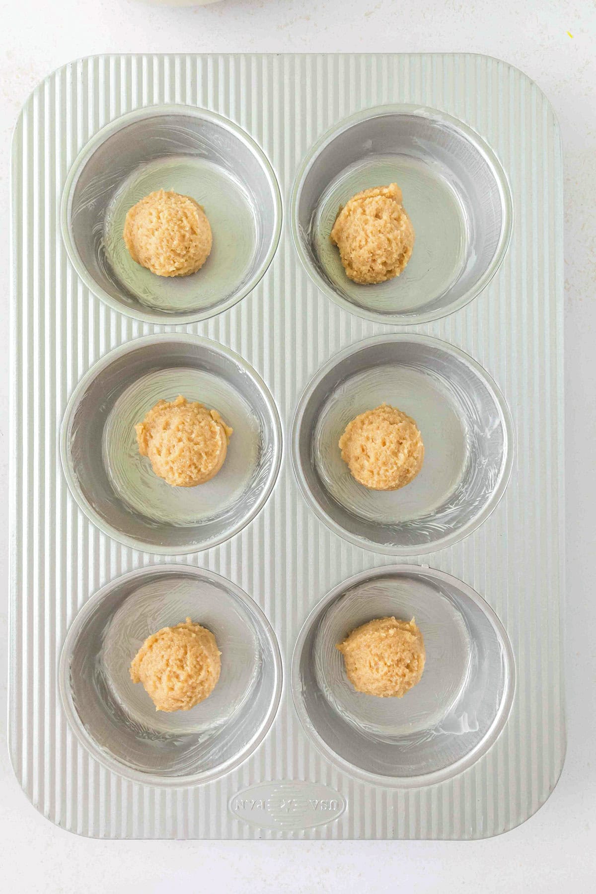 Six unbaked Pineapple Upside Down Sugar Cookie dough portions are spaced evenly in a greased metal muffin pan with six wells, viewed from above on a white surface.