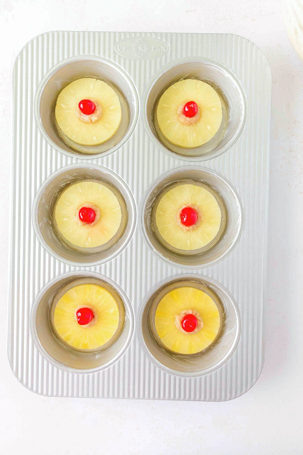 Six muffin tin cups each contain a pineapple ring with a maraschino cherry in the center, arranged neatly in a metal baking pan on a white surface, ready to be transformed into delightful Pineapple Upside Down Sugar Cookies.