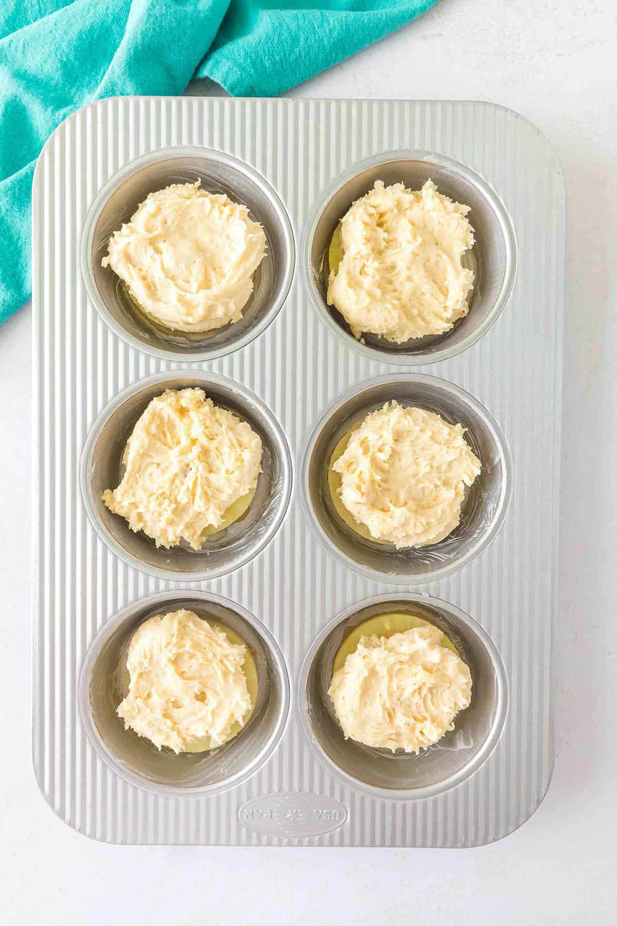 Six muffin tin cups filled with unbaked Pineapple Upside Down Sugar Cookies batter, set on a white countertop with part of a teal cloth visible in the corner.