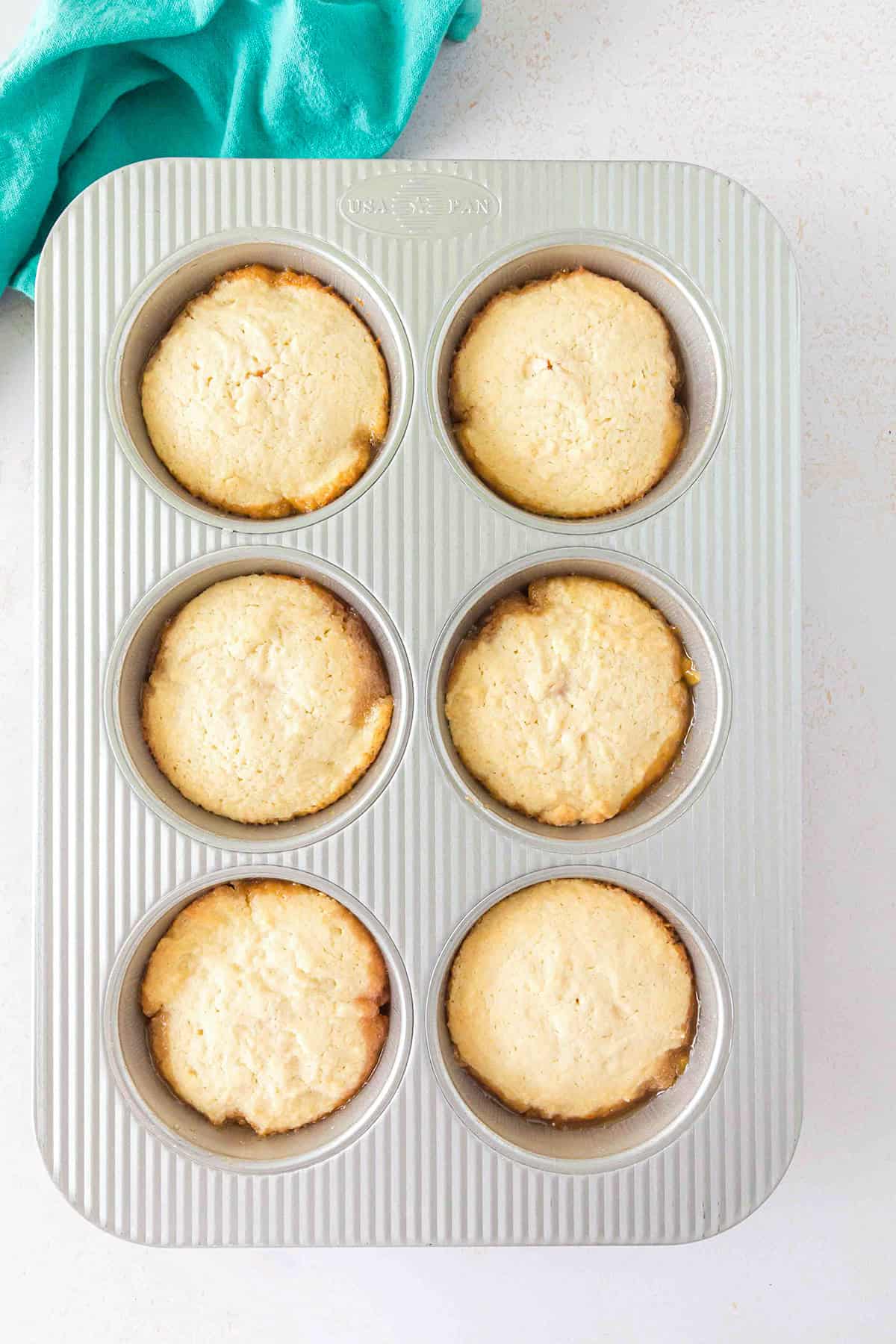 Six baked muffins in a silver muffin pan sit on a light-colored surface with a teal cloth in the upper left corner, reminiscent of the golden tones found in Pineapple Upside Down Sugar Cookies.